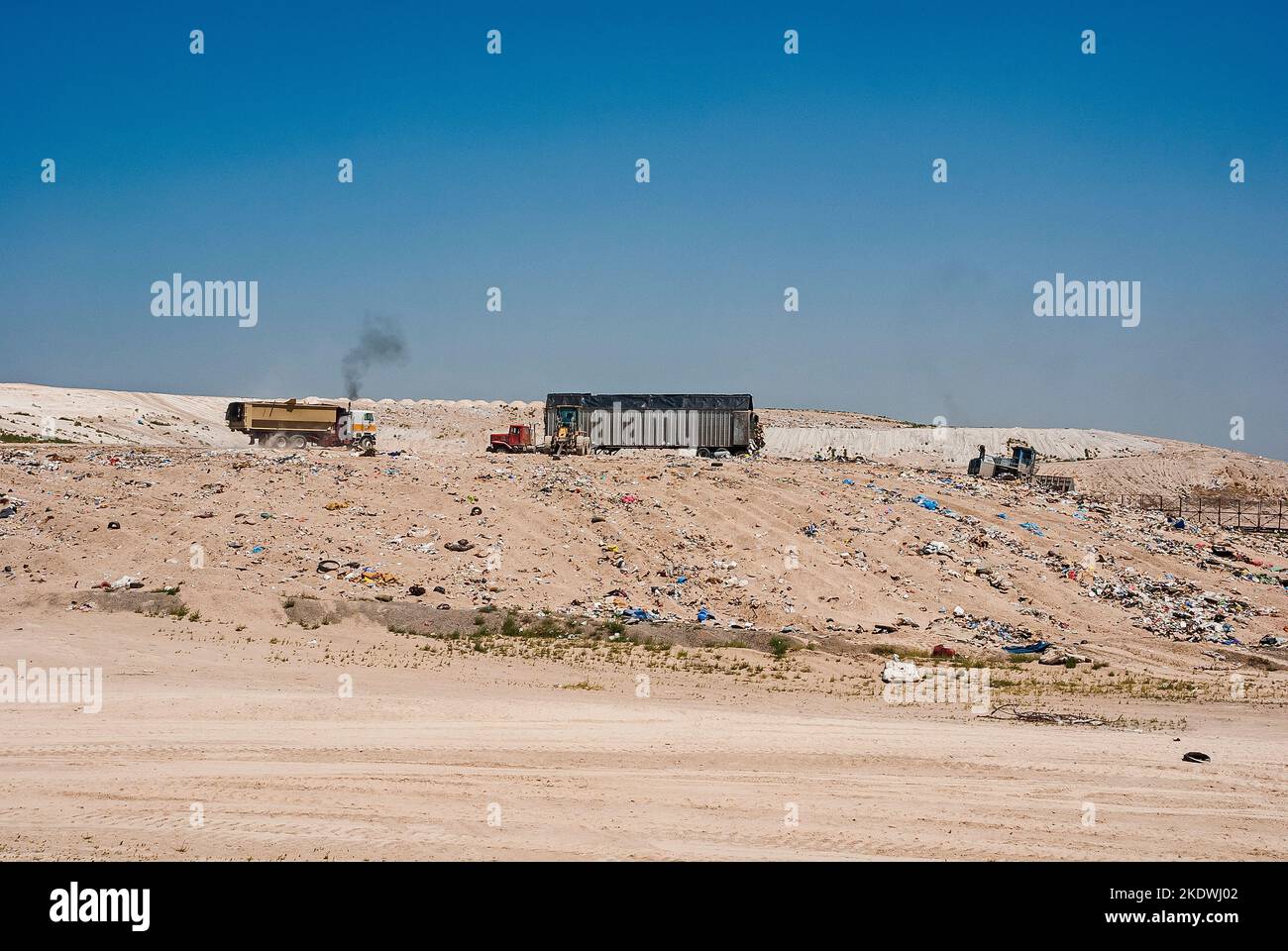 A tractor-trailer semi and two soil compactors on flat land in an active landfill.  One truck is emitting diesel smoke. Stock Photo