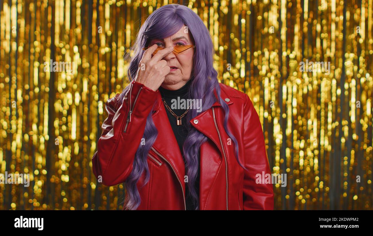 I am watching you. Woman with purple hairstyle wig pointing at his eyes and camera, show I am watching you gesture, spying on someone. Senior elderly grandmother on shiny disco party foil background Stock Photo