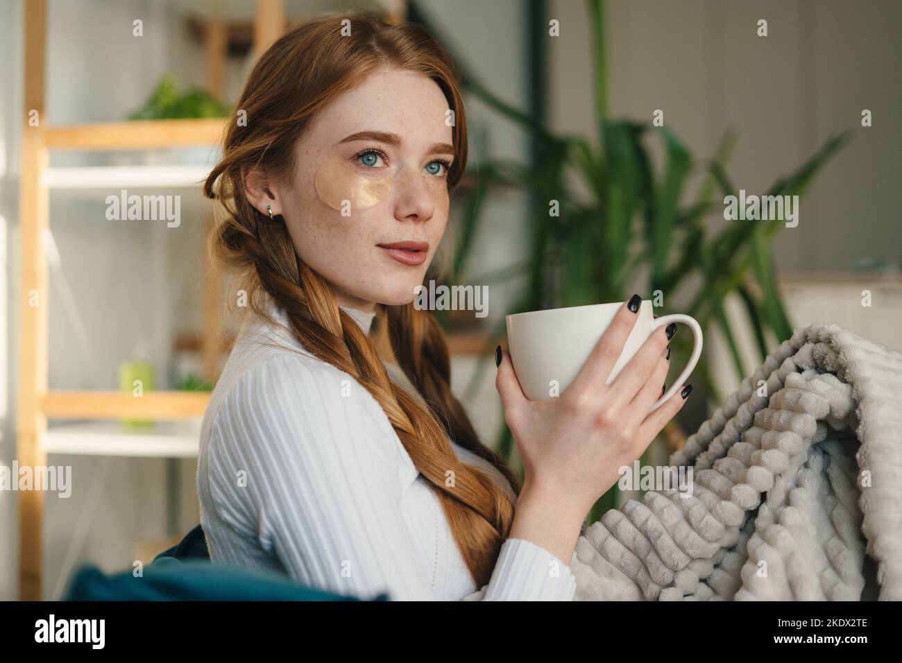 Young beautiful freckled woman with eye patches drinking morning coffee on sofa. Morning habits and home beauty routine. Beauty treatment. Female Stock Photo