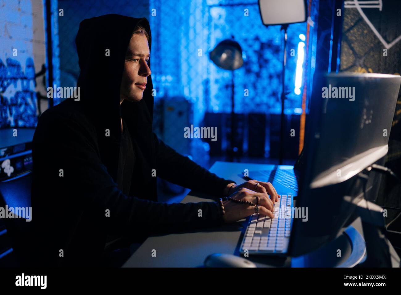 Side view of hidden hacker man wearing sweatshirt with hood engaged in hacking into security systems, sitting in dark room with blue neon lights Stock Photo