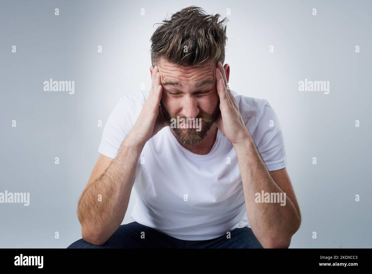 Does anyone have a headache tablet. a young man with a headache holding his head while sitting on a chair in the studio. Stock Photo