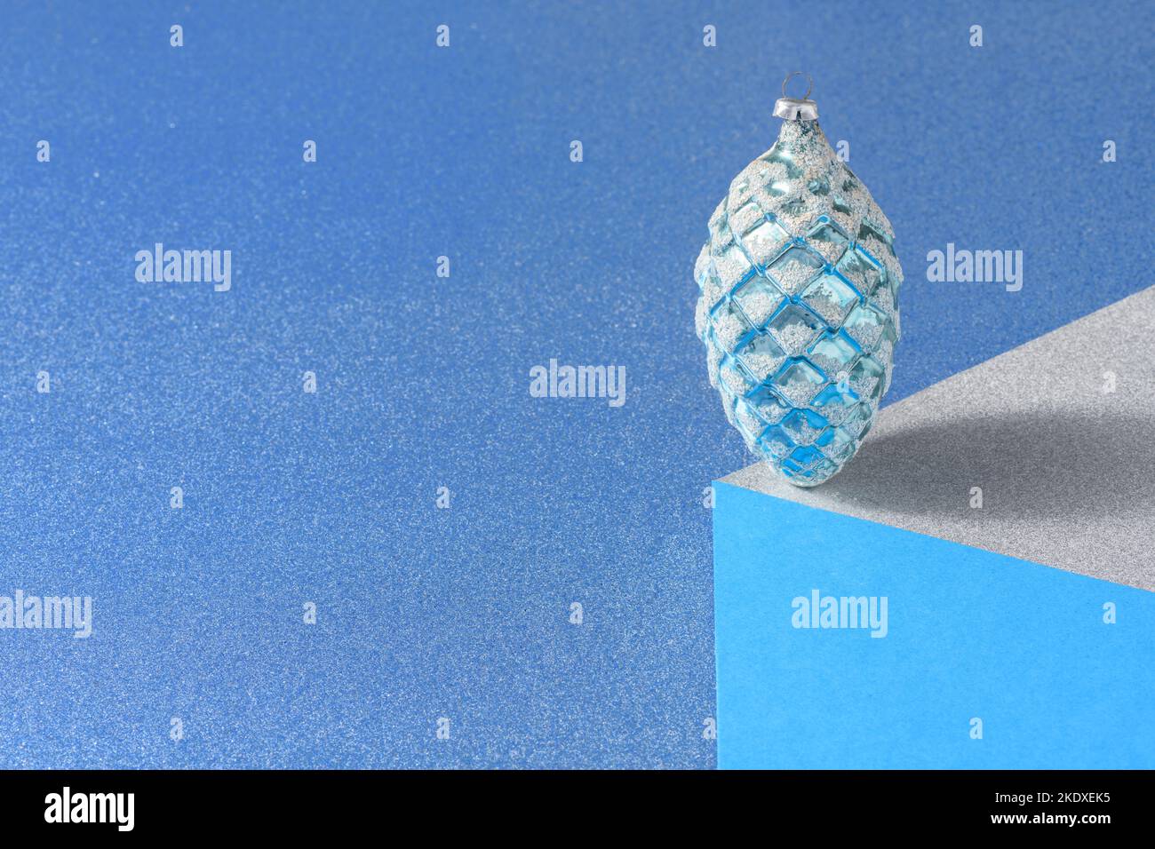 Optical illusion of a vintage christmas bauble in the shape of a pine cone standing in balance on a paper cube against a blue background. Stock Photo