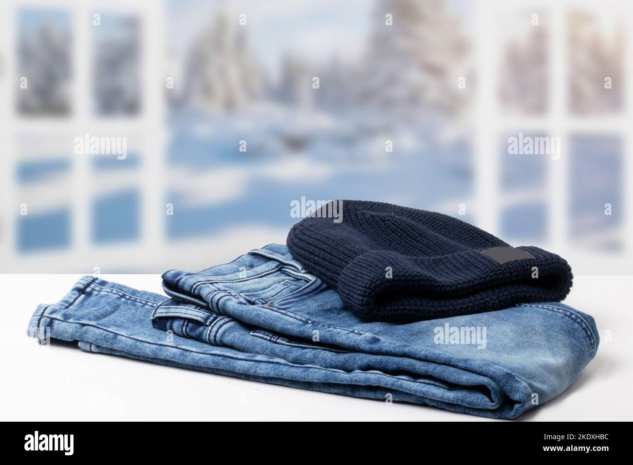 Autumn clothes. Fashionable folded jeans and a knitted blue Rapper, Beanie or Baseball hat or cap on table over abstract blurred winter landscape. Cop Stock Photo