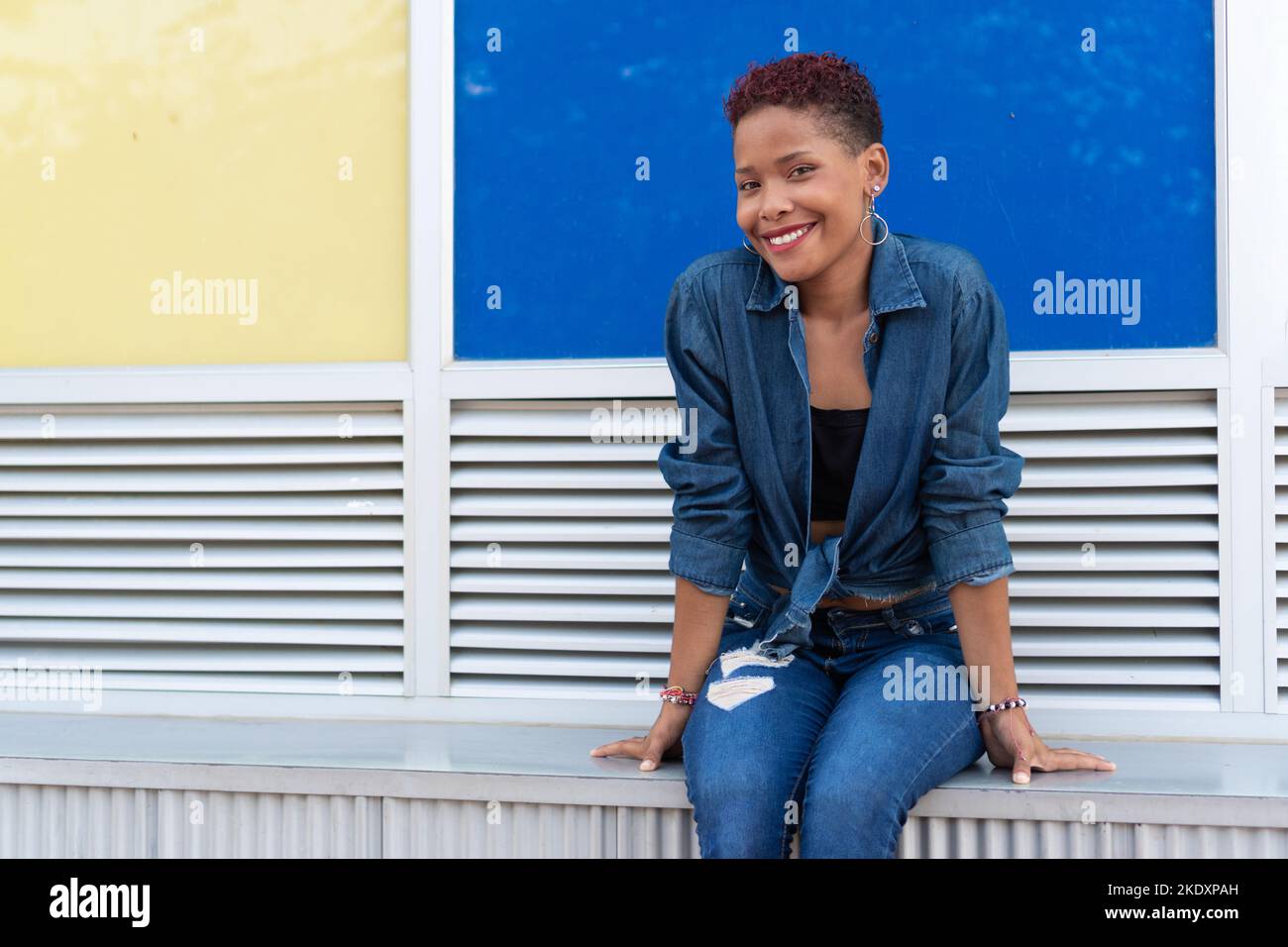 Positive Colombian female in casual clothes with short hair smiling and  looking at camera while sitting on bench against colorful wall on city  street Stock Photo - Alamy