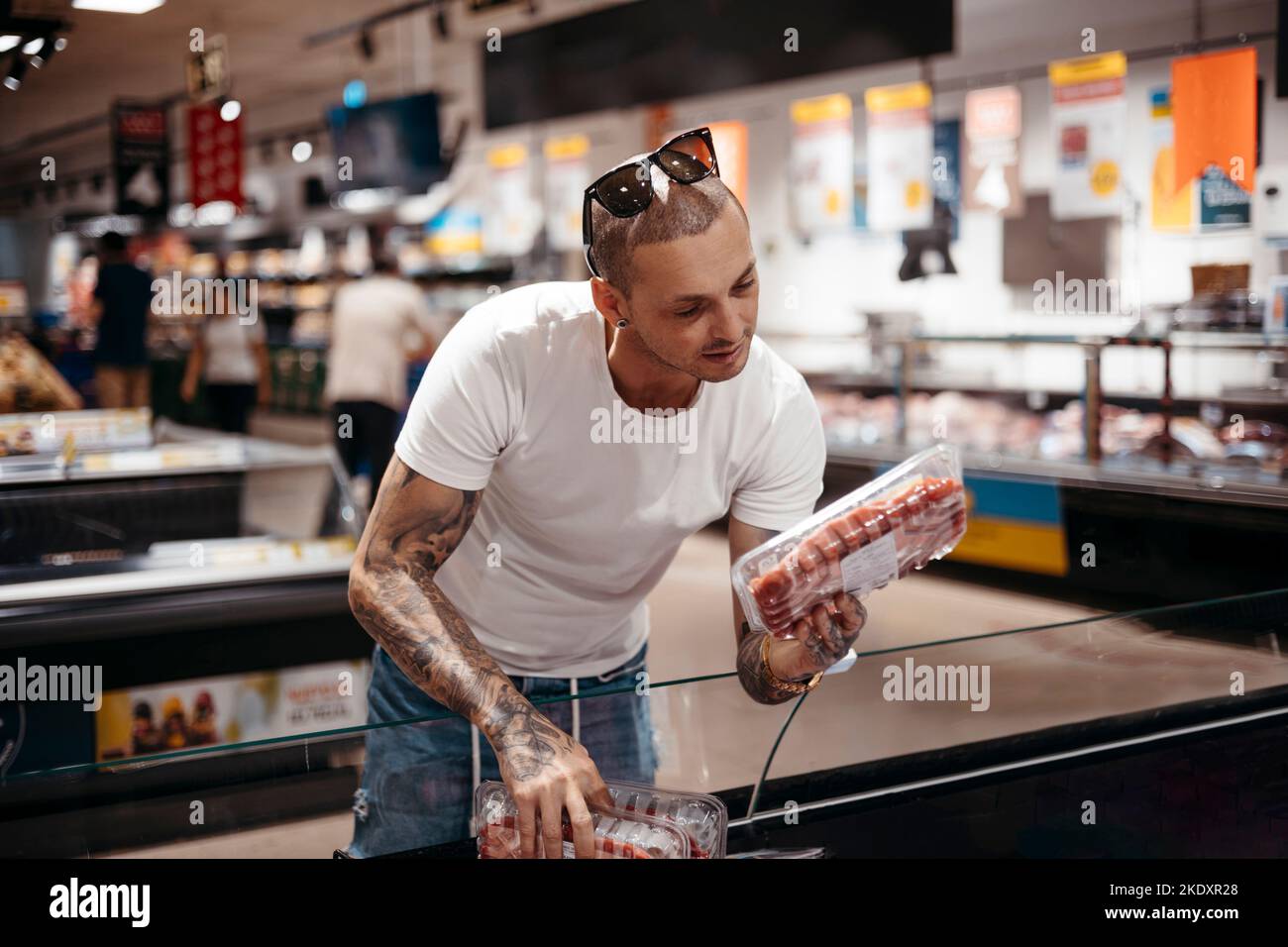 Tattooed male customer in casual clothes standing near chest freezer and picking steak for dinner in supermarket Stock Photo