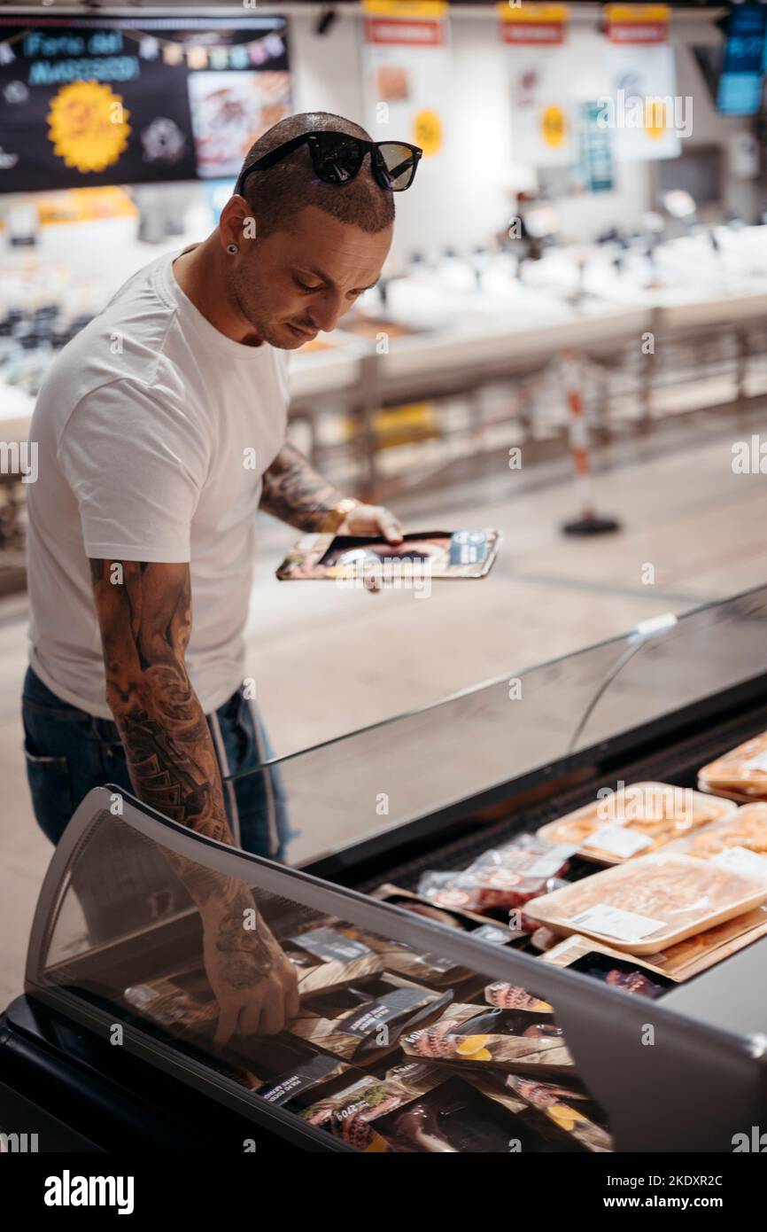 Tattooed male customer in casual clothes standing near chest freezer and picking steak for dinner in supermarket Stock Photo
