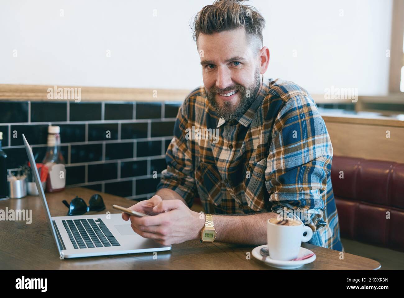 Caffeine and connectivity. Portrait of a young man using his cellphone and laptop while sitting in a cafe. Stock Photo