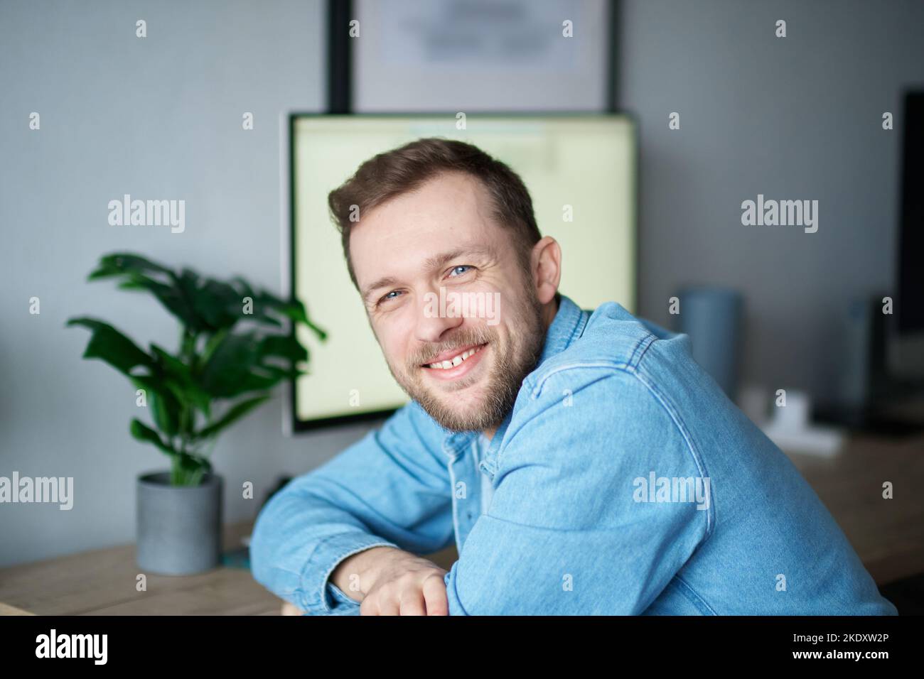 Cute attractive caucasian man in denim shirt with positive emotion and computer on background. Cheerful bearded male freelancer sitting in home office looking at camera smiling. High quality image Stock Photo