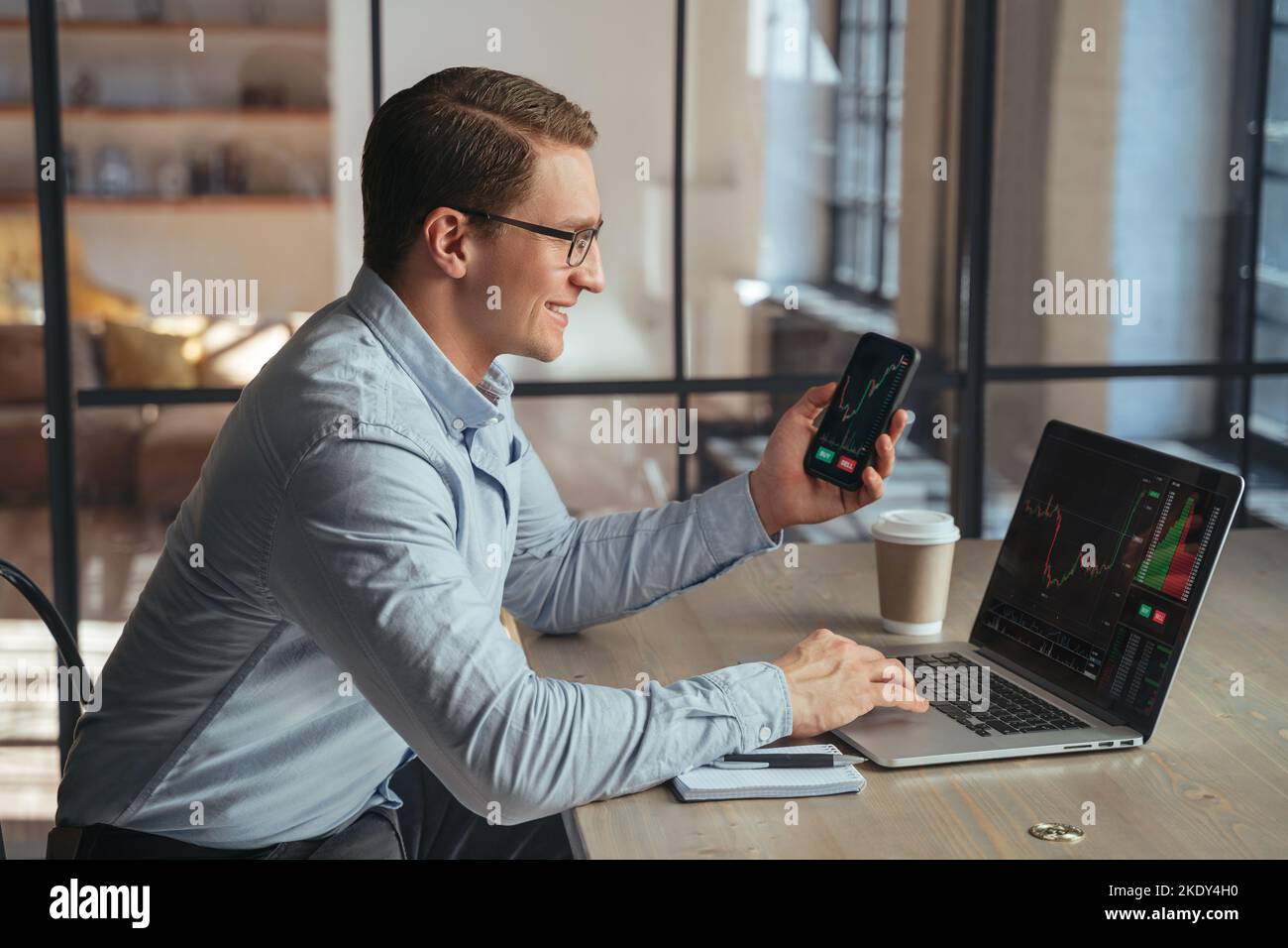 Smiling businessman crypto trader holding smartphone checking candlestick graph on laptop, buying and selling currency, happy to notice tendency of btc rising up, golden bitcoin near on table Stock Photo