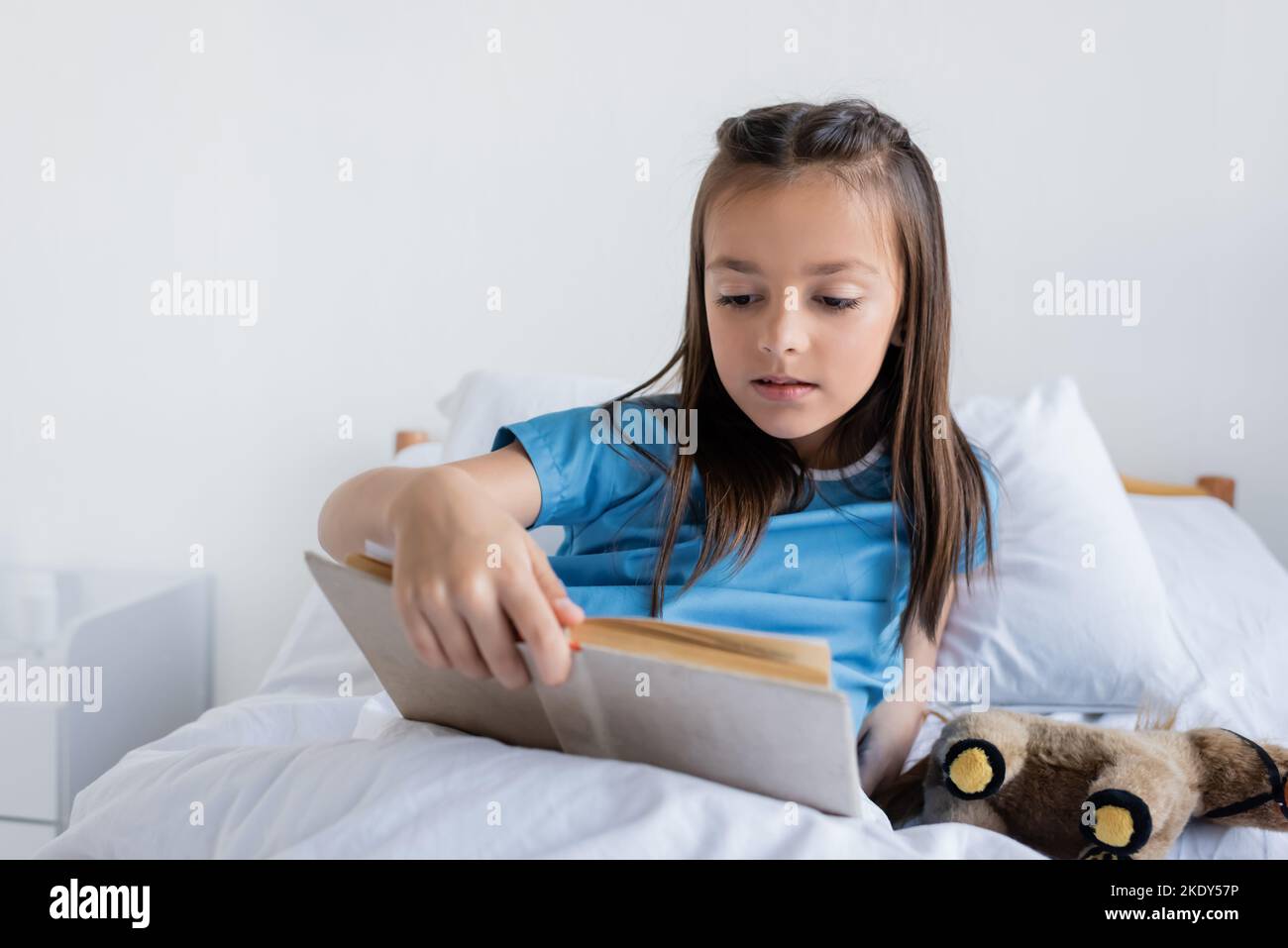 Girl in patient gown reading book on bed in hospital ward,stock image