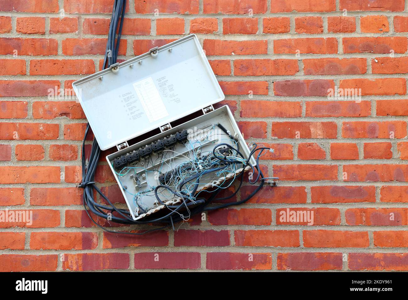An outdoor telephone box with tangled wires and corrosion, haphazardly dangling off a brick wall. Stock Photo