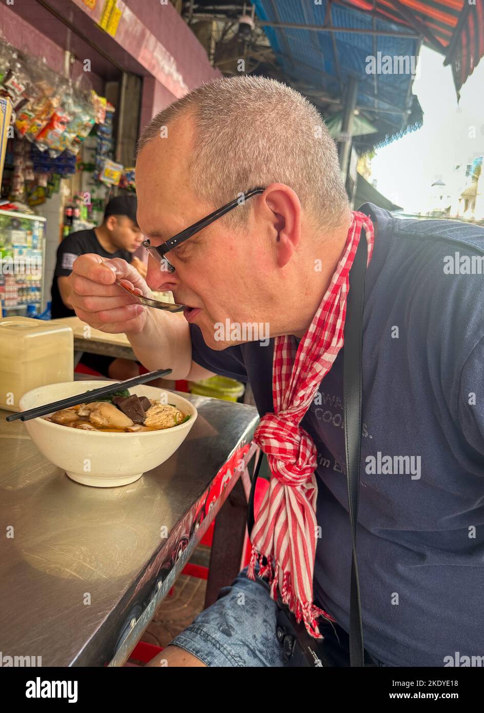 A middle-aged foreign tourist eats a bowl of bun rieu, a form of Vietnamese noodle soup, in central Phnom Penh, Cambodia. Stock Photo