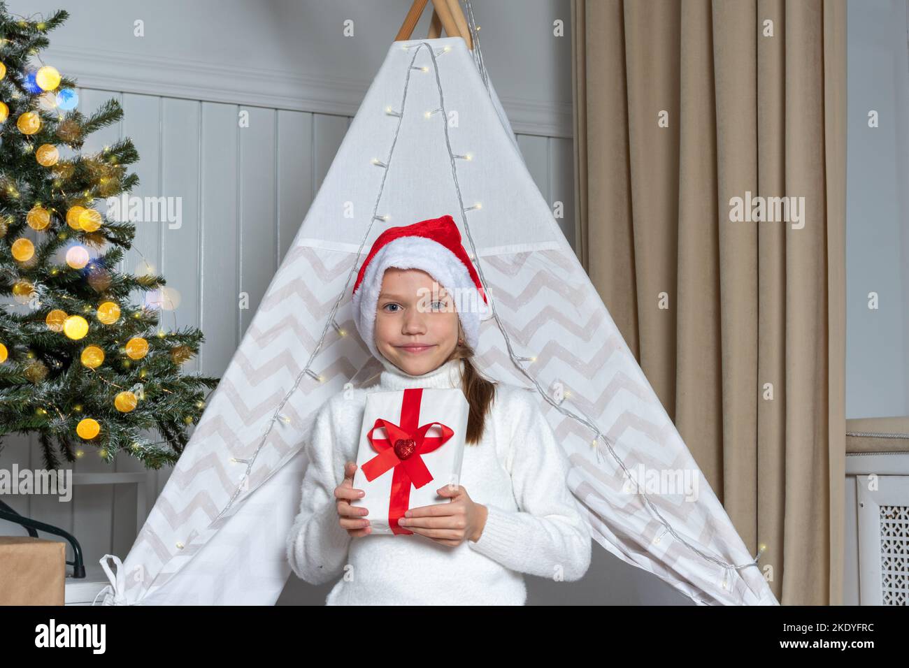 A pretty girl in a Santa hat holds her gifts for Christmas New Year lying in a children's room decorated with garlands. Christmas gifts for children. Stock Photo