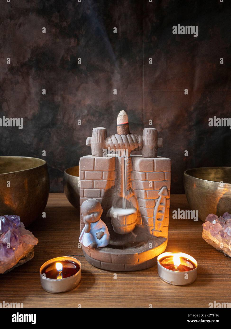 Still life with incense burner with cascade of reflux incense, candles, druses and Tibetan bowls, on wooden table and dark background. Stock Photo