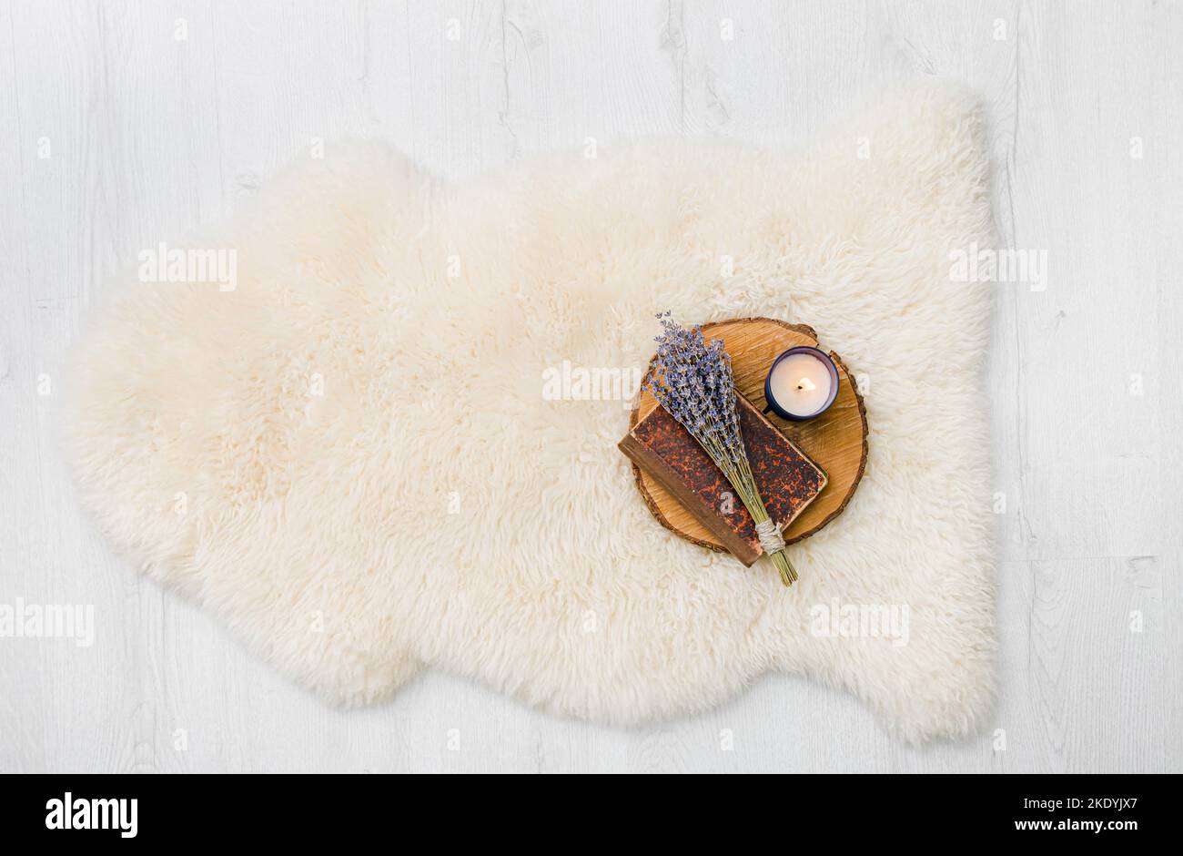 Flat lay view of white warm and cozy whole real decorative sheepskin rug on white wooden board floor, on top is book, flowers and candle. Copy space. Stock Photo