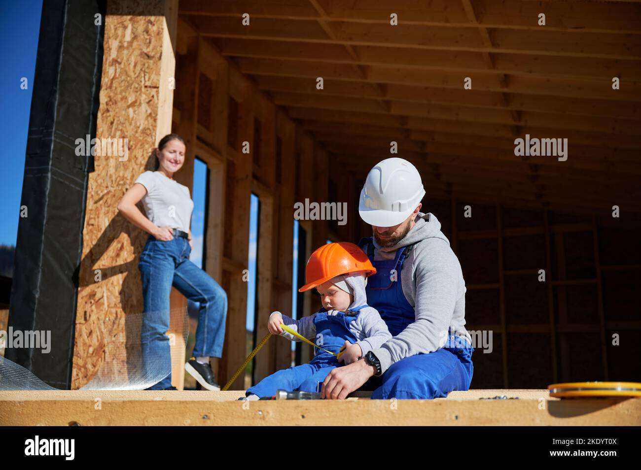 Father, mother and son building wooden frame house in the Scandinavian style barnhouse. Toddler boy helping his daddy, while woman looking for them on construction site. Carpentry, family concept. Stock Photo