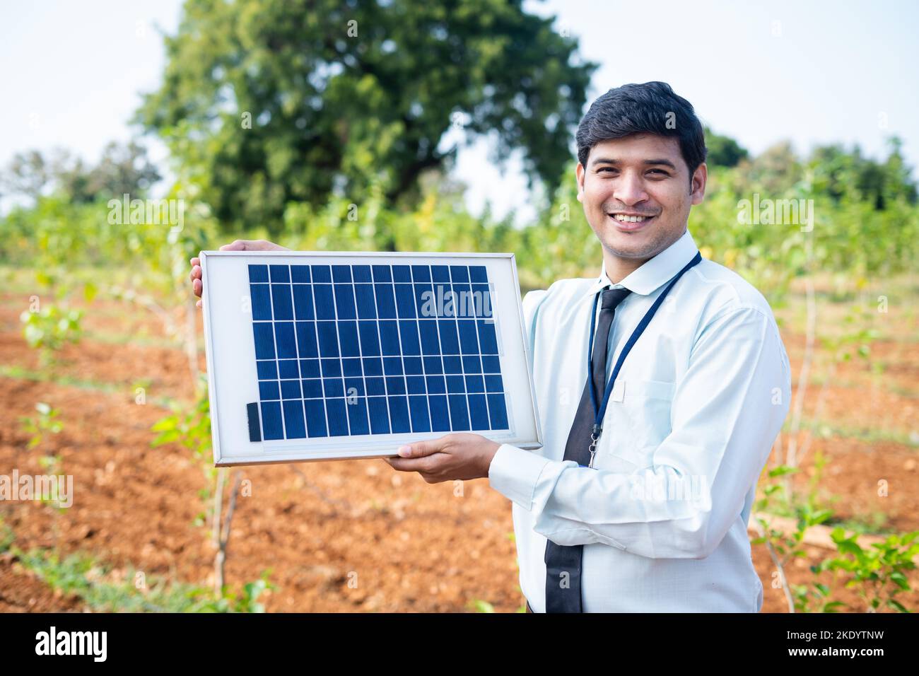 Confident banking officer showing solar panel by looking at camera at agricultural land - concept of advertisement, recommending, renewable electric Stock Photo