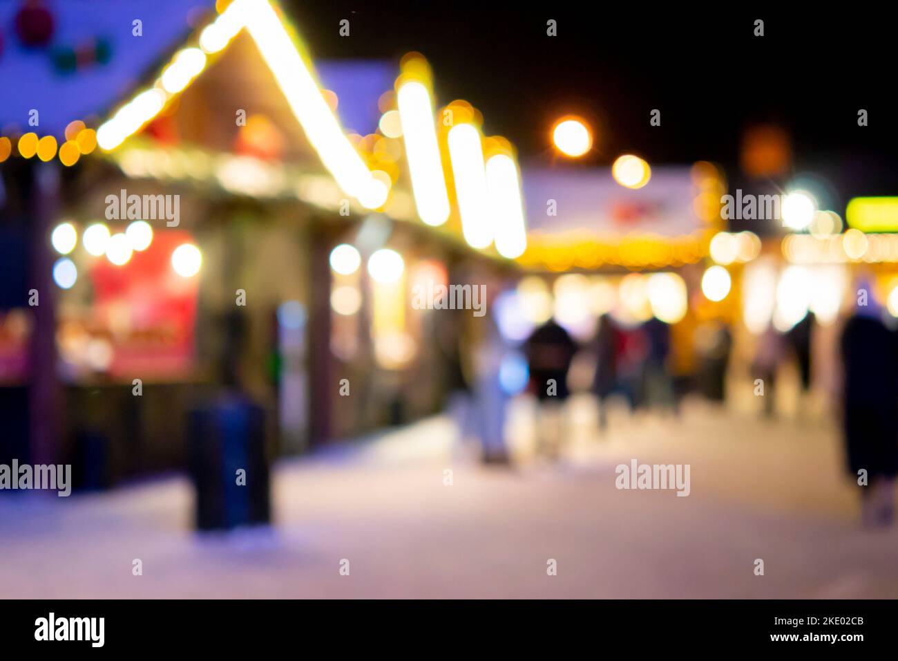 Blurred background. People walk in city square on winter night. Black silhouettes of people walking near houses decorated luminous illumination. White light bokeh blur spots from glowing house lights Stock Photo
