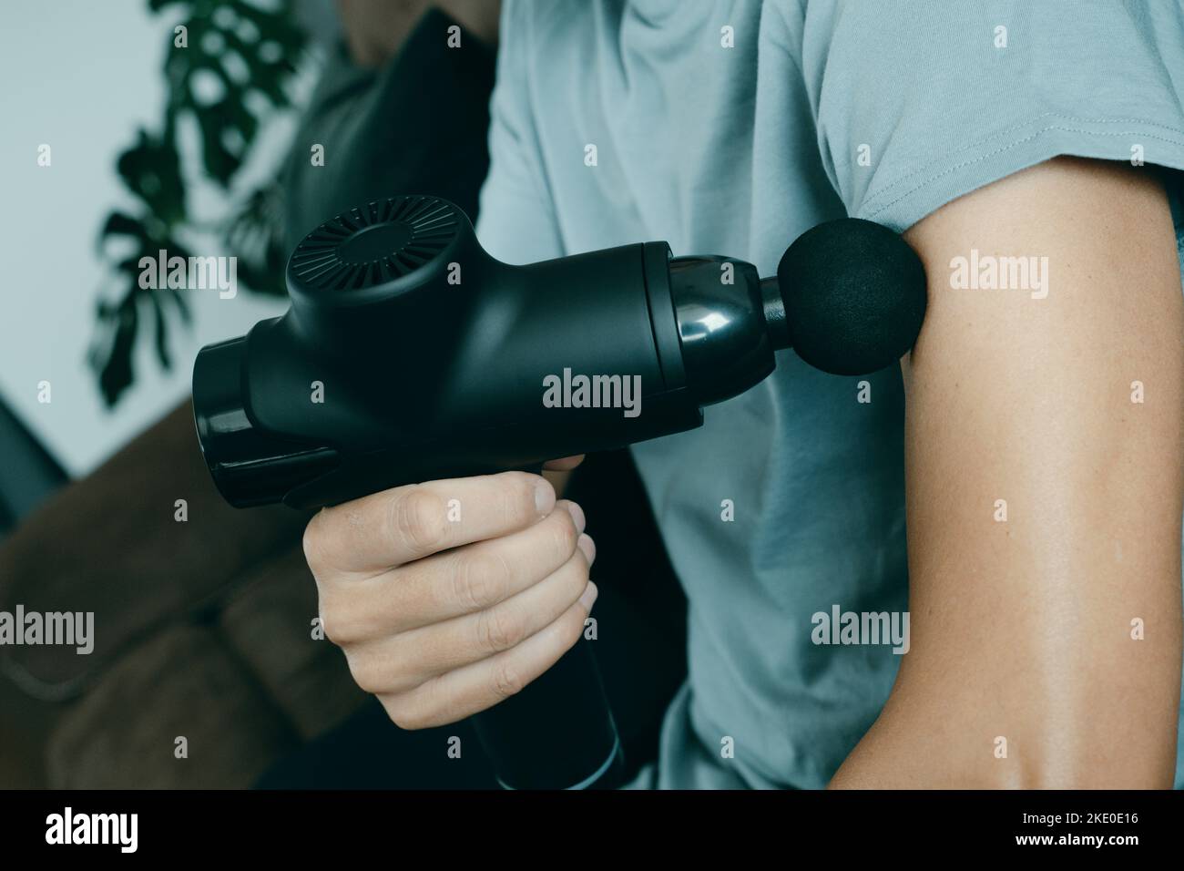a young caucasian man at home uses a massage gun to massage the muscles of his arm, sitting on the couch of his living room Stock Photo