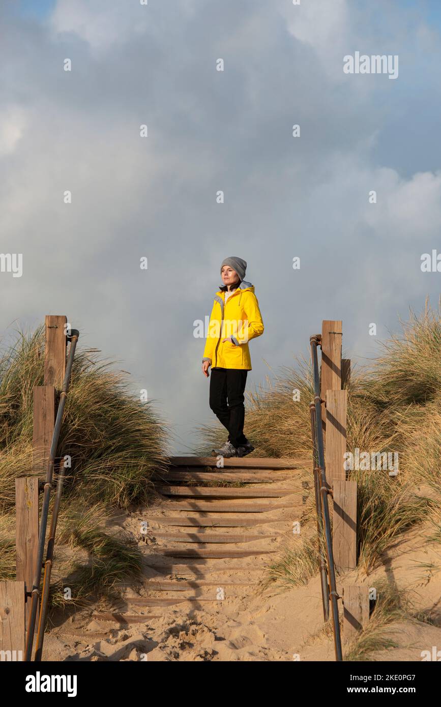 Woman wearing a yellow coat standing at top of sand dunes, looking at the view. Stock Photo