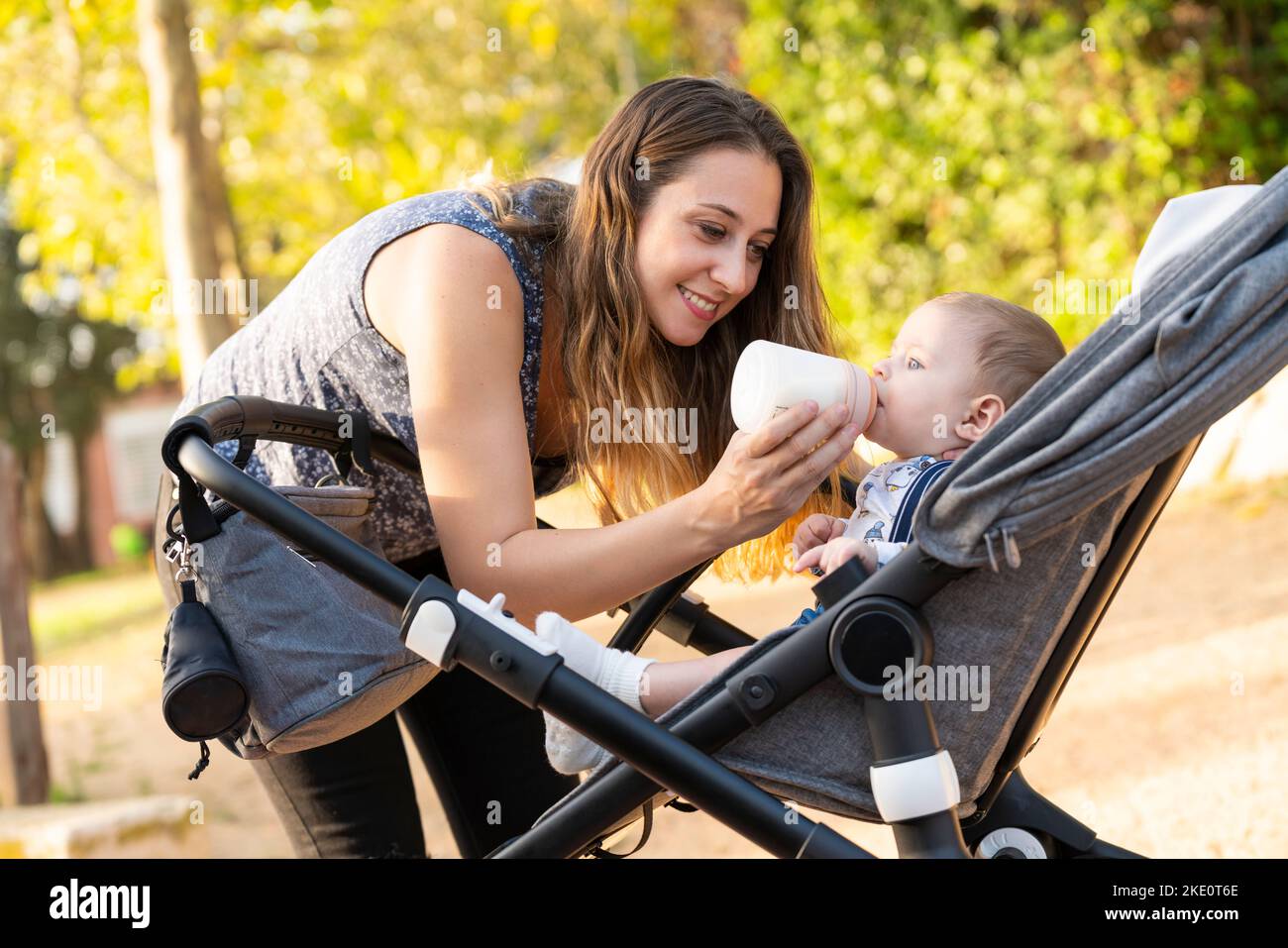 Mother feeding her son with a milk bottle in the park inside the baby stroller Stock Photo