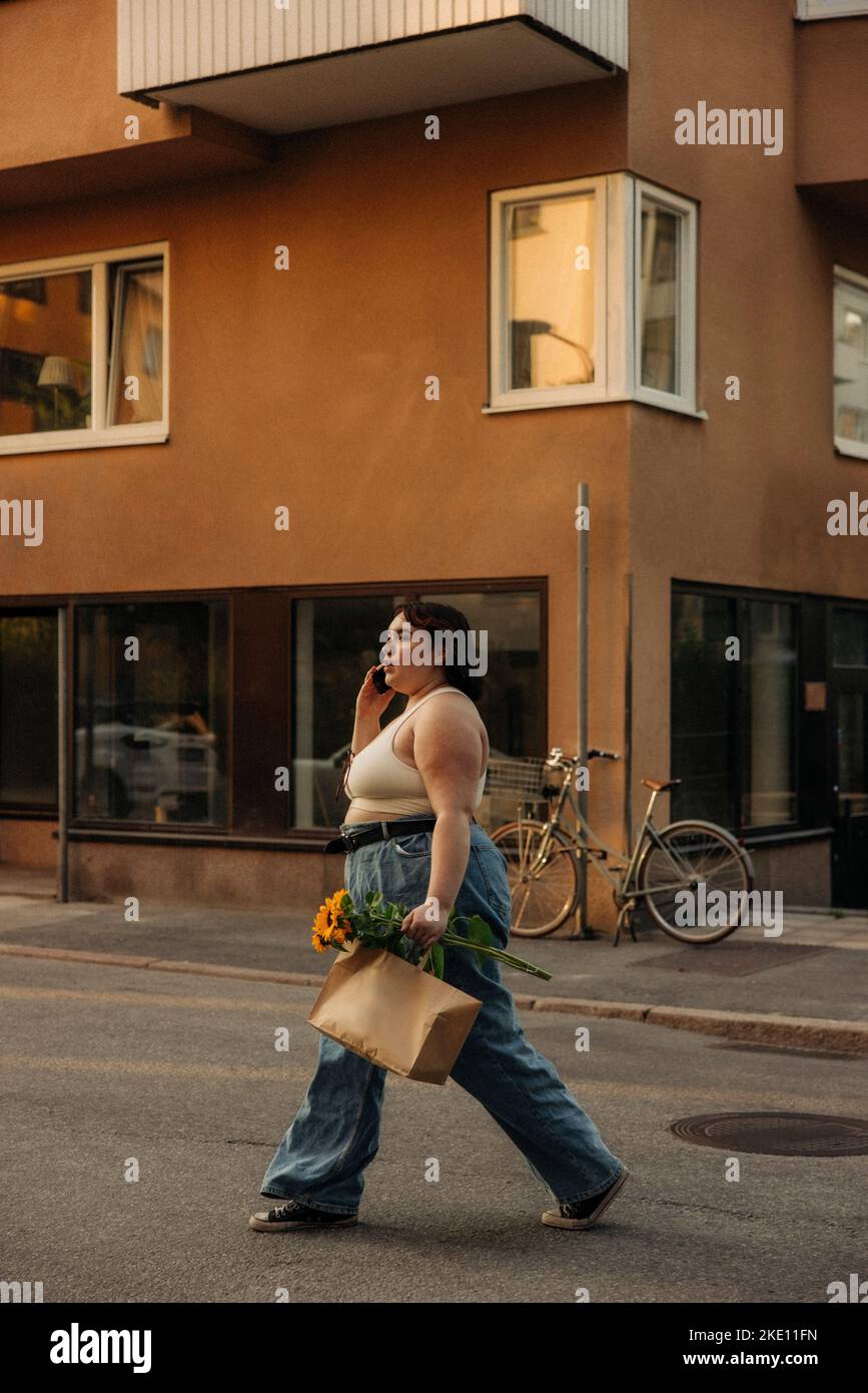 Side view of voluptuous woman talking on mobile phone while walking with shopping bag on road Stock Photo