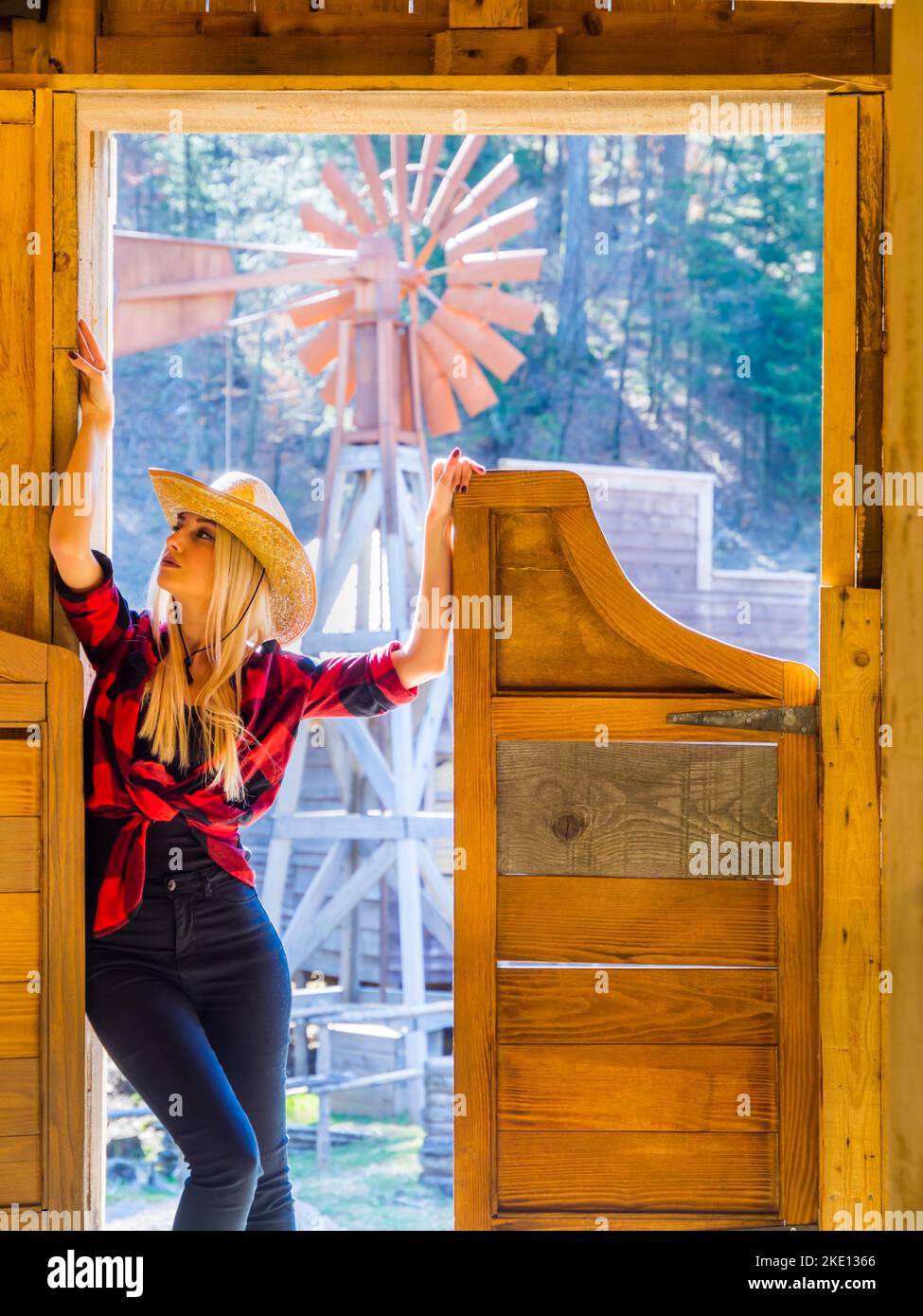 Young woman American western clothing style with plaid shirt and stetson hat standing on entrance of saloon bar looking away aside up Stock Photo