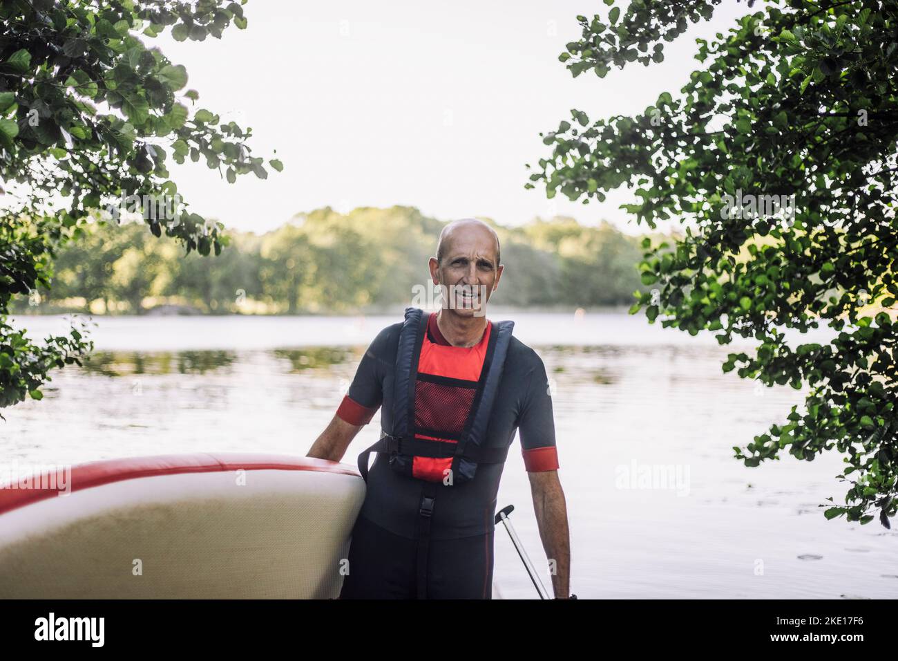Mature man wearing wetsuit and life jacket while holding paddleboard