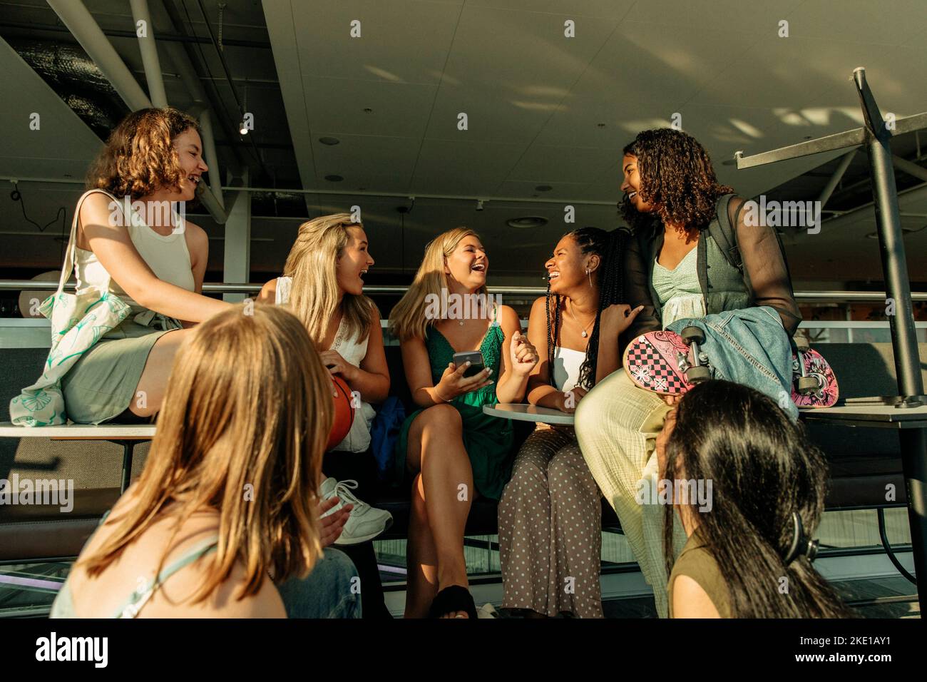 Cheerful teenage girls talking with each other while sitting outside mall during sunny day Stock Photo