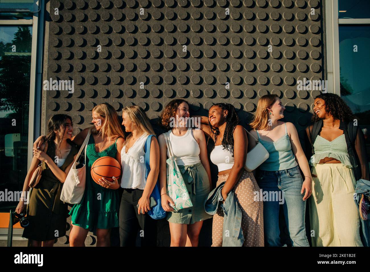 Happy teenage girls looking at each other while standing against wall during sunny day Stock Photo