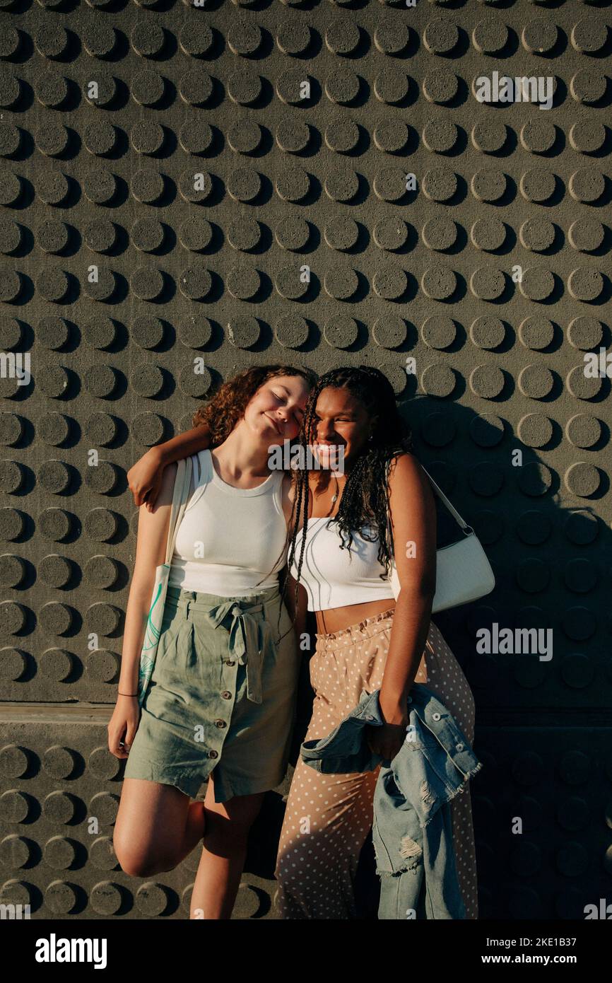 Portrait of happy teenage girl standing with arm around friend against wall during sunny day Stock Photo