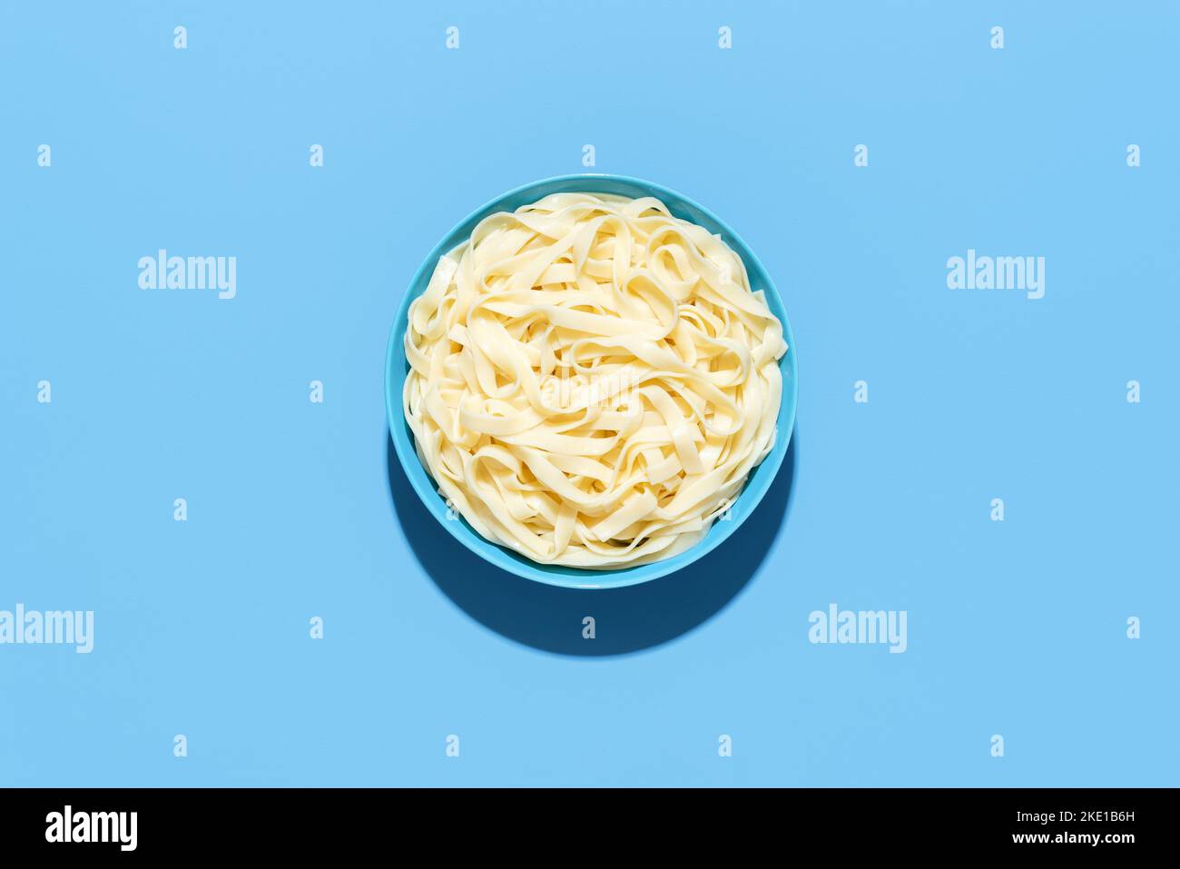 Above view with a bowl full of tagliatelle pasta, minimalist on a blue table. Boiled pasta without sauce in bright light on a colorful background Stock Photo