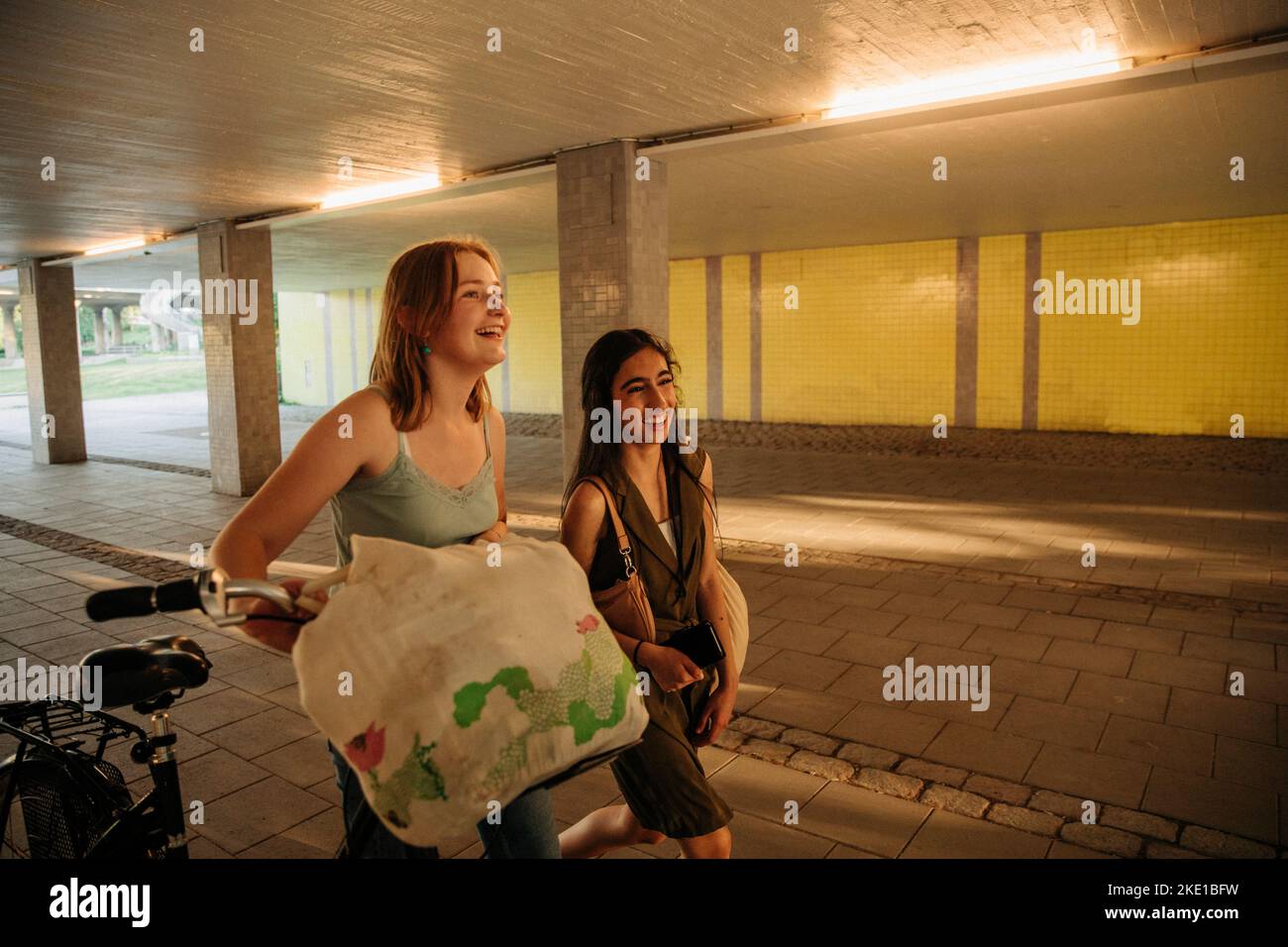 Smiling teenage girl walking with bicycle by female friend under bridge Stock Photo