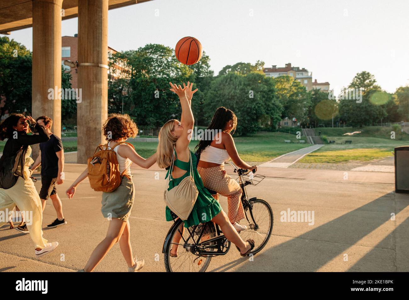 Rear view of teenage girl throwing basketball while sitting with female friend riding bicycle at park Stock Photo