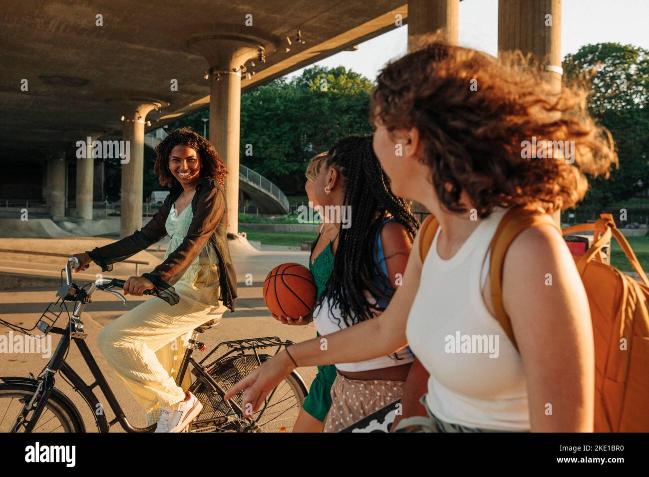 Teenage girls looking at smiling female friend riding bicycle under bridge Stock Photo