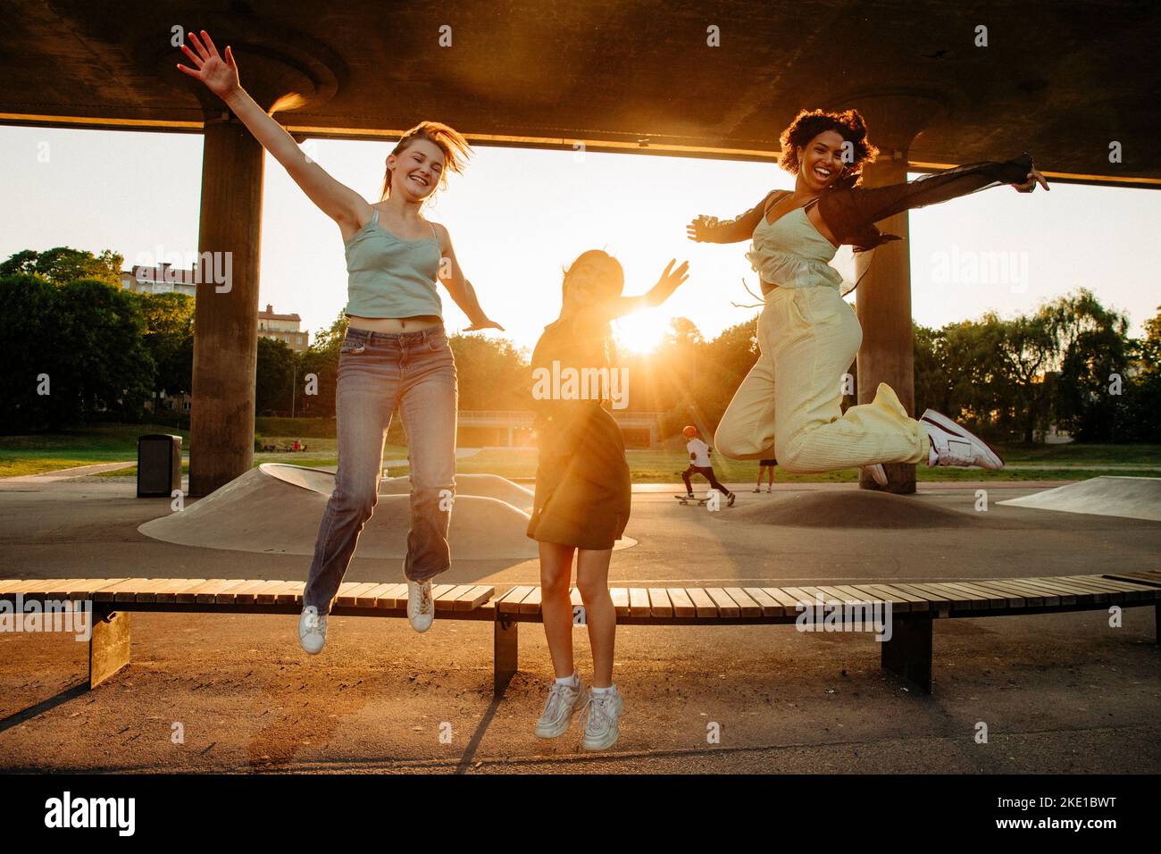 Portrait of happy teenage girls with arms outstretched jumping by bench during sunset Stock Photo