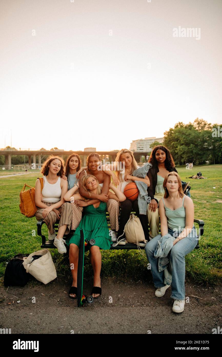 Multiracial teenage girls sitting on bench at park during sunset Stock Photo