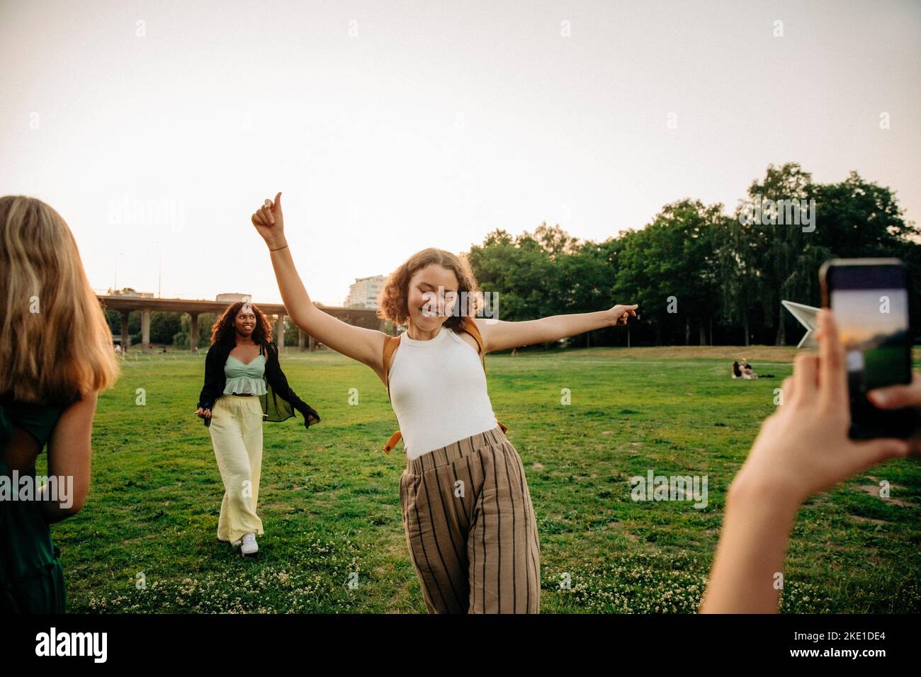 Teenage girl photographing carefree female friend dancing at park during sunset Stock Photo