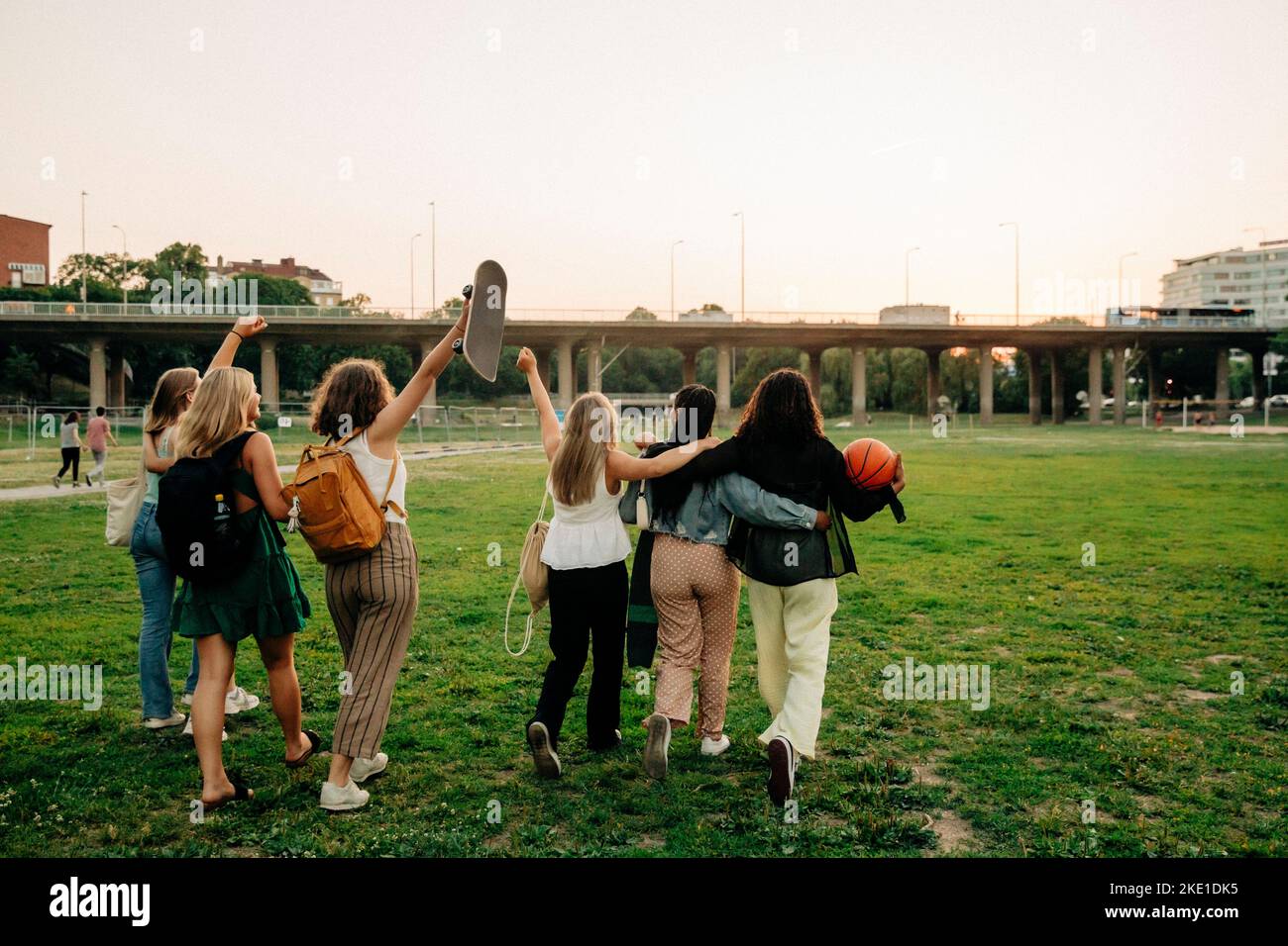 Rear view of teenage girls with hand raised walking by friends at park Stock Photo