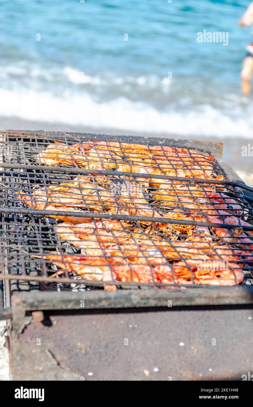 Grilled shrimp in a barbecue grill, selective focus on foreground with shallow depth of field with blurred beach pebbles and a wave of turquoise color Stock Photo