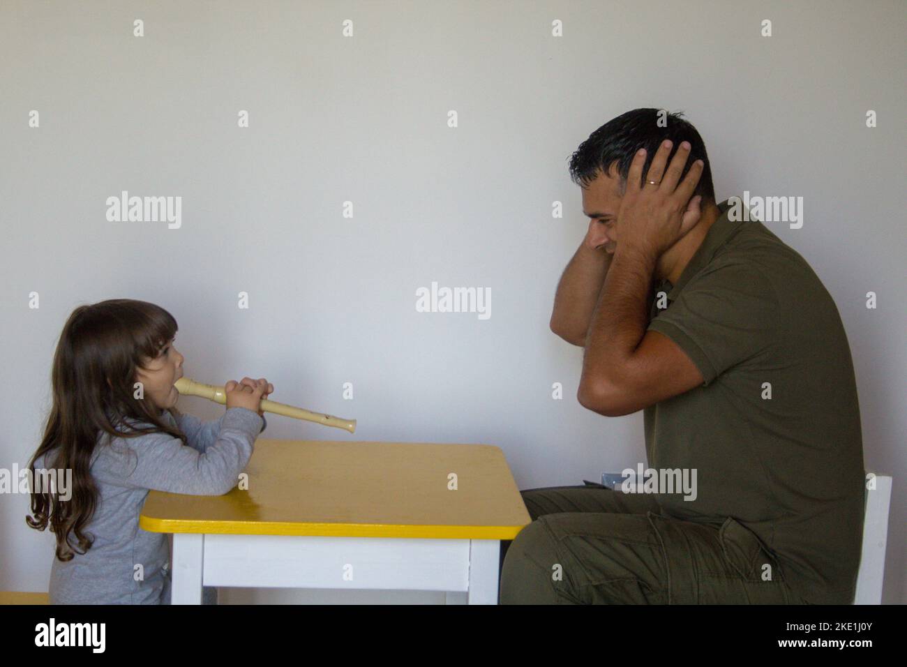 Little girl playing the flute and dad plugging his ears. Music lessons of a little girl learning to play. Stock Photo