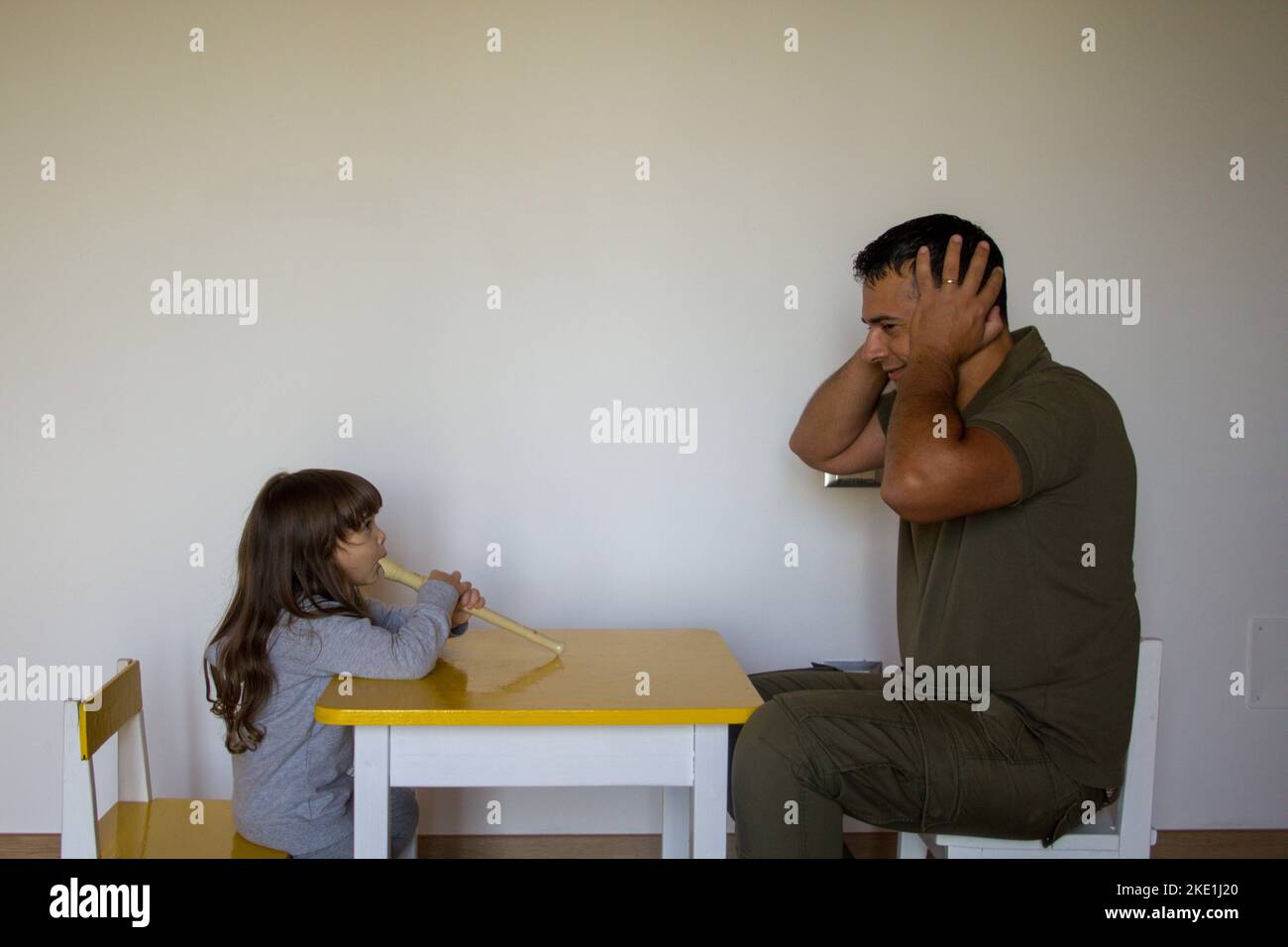 Little girl playing the flute and dad plugging his ears. Music lessons of a little girl learning to play. Stock Photo