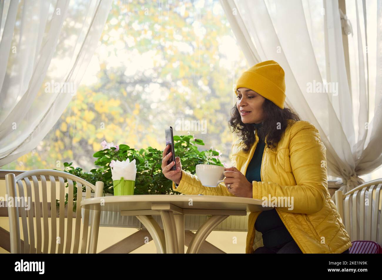 A body positive pretty woman in yellow jacket, sitting at table with coffee, having nline conference on her mobile phone Stock Photo