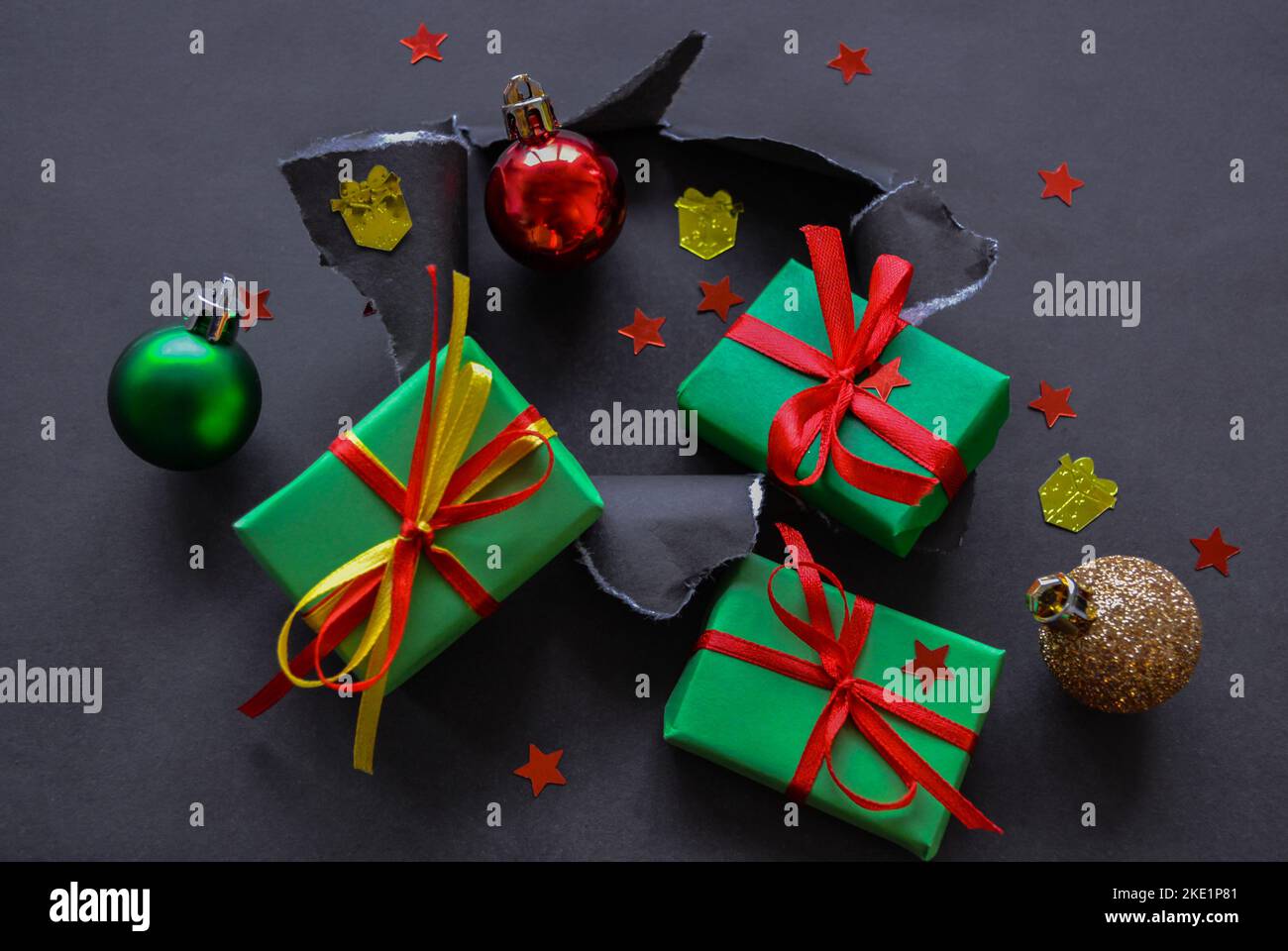 Gifts in green paper with red and yellow ribbons, christmas balls and confetti in the form of red stars and gold presents fall out a torn hole in the Stock Photo