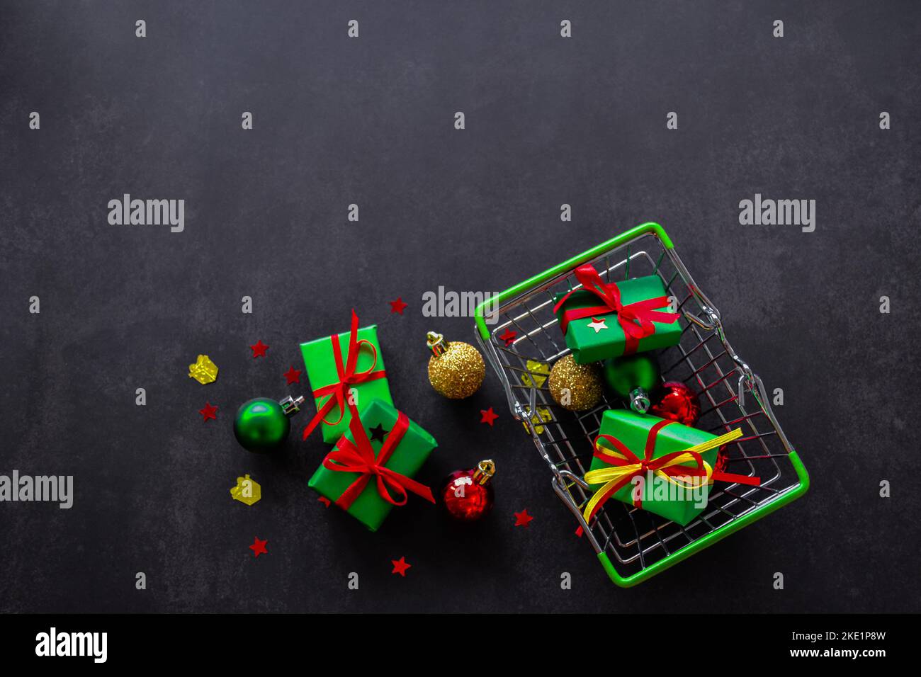 Top view of shopping basket with gifts in green paper, red and yellow bows, Christmas balls of red, green, gold on a black background. Some gifts, bal Stock Photo