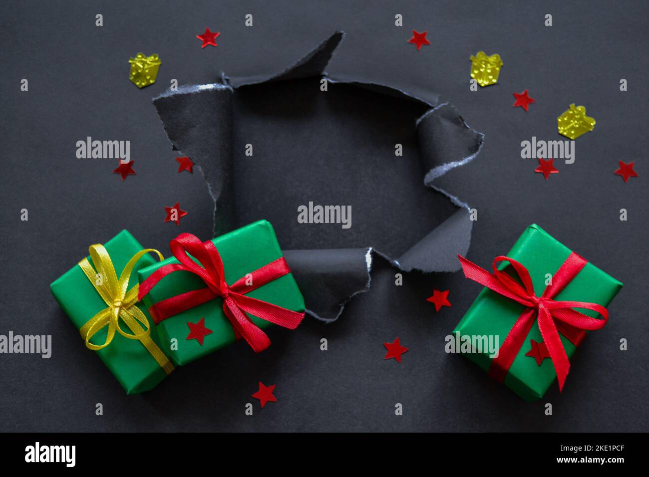 Gifts in green paper with red and yellow ribbons and confetti in the form of red stars and gold presents fall out a torn hole in the black paper. Big Stock Photo