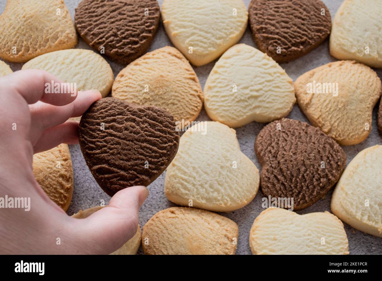 Hand holds a heart-shaped chocolate cookie against a backdrop of many colorful cookies. Love concept, homemade and food background. Stock Photo