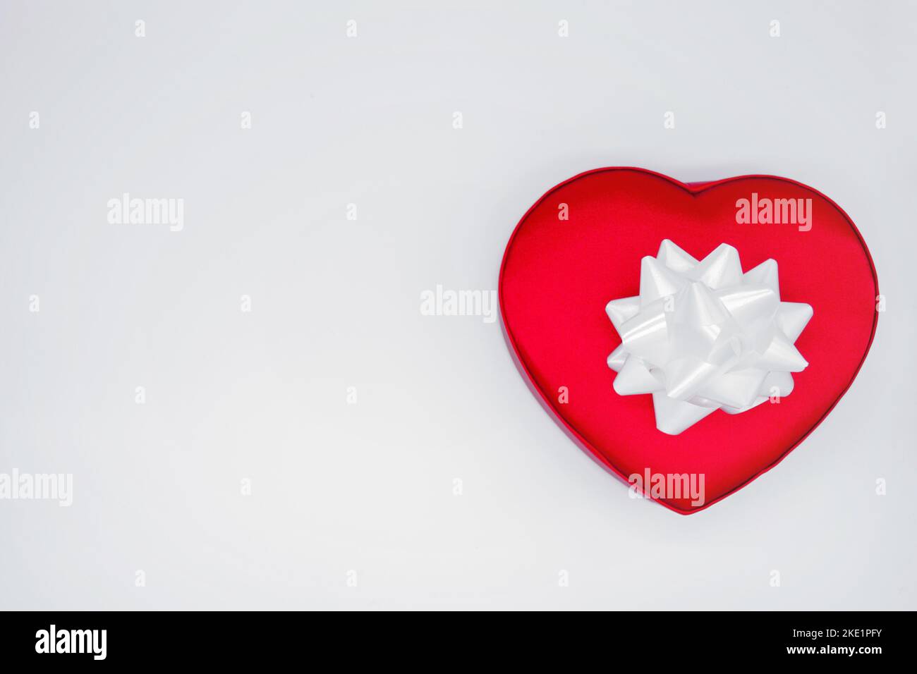 Red satin box in shape of a heart, tied with a with large white flower bow on a white background. Concept of love, Valentine's day, gift, surprise. Stock Photo