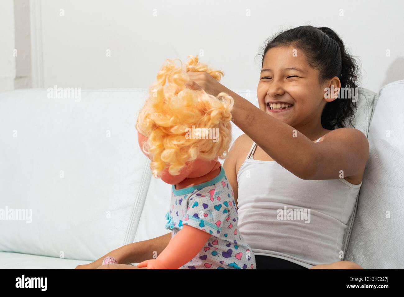 latina girl with a big smile, playing with her doll on the sofa. brunette combing her toy. Stock Photo