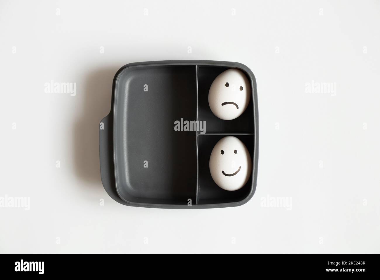 Two eggs with a joyful smile and a sad one lie in a plastic container on a white background, a choice of joy and sadness, lunch Stock Photo